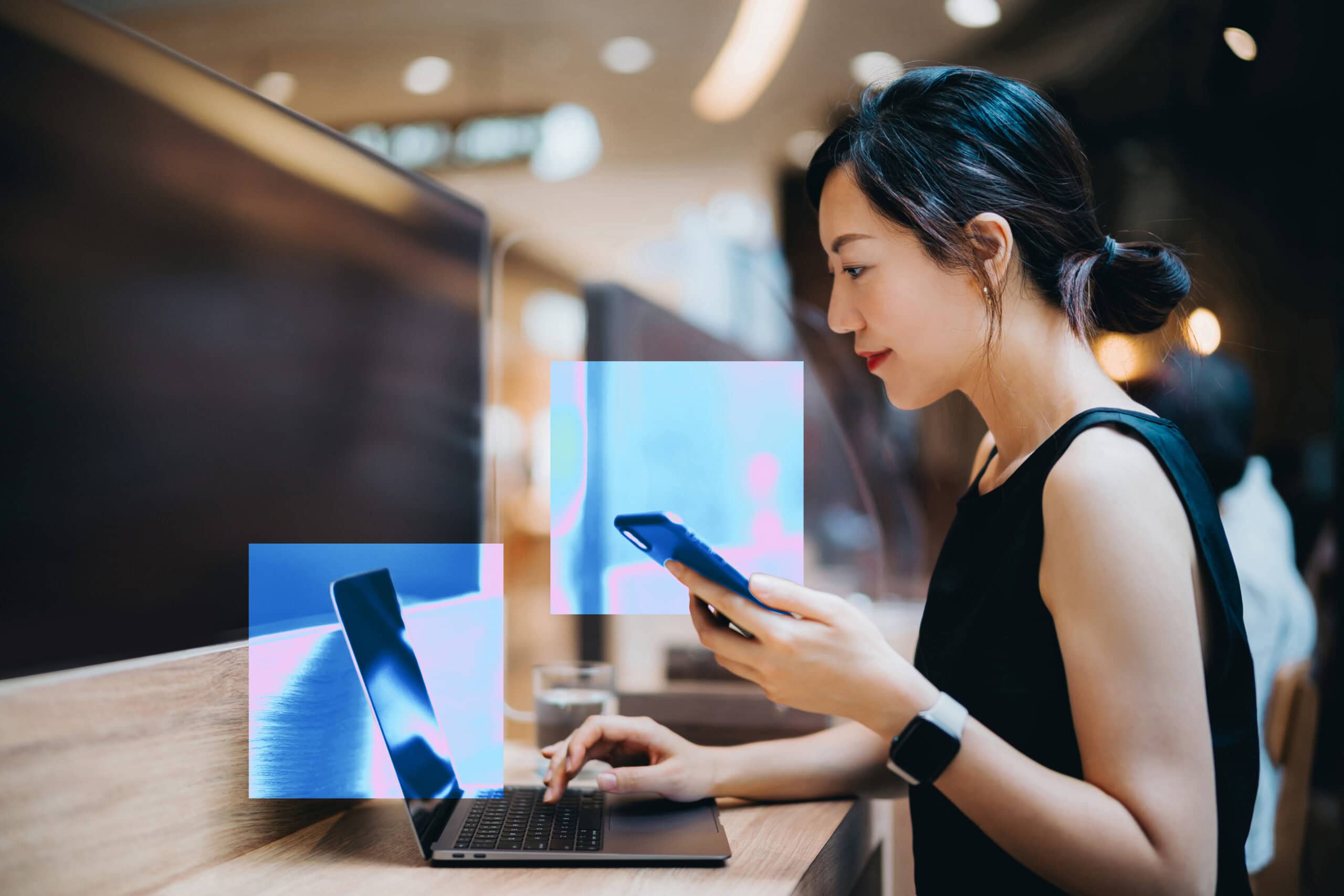 A woman looks at her computer screen and holds a phone in her left hand